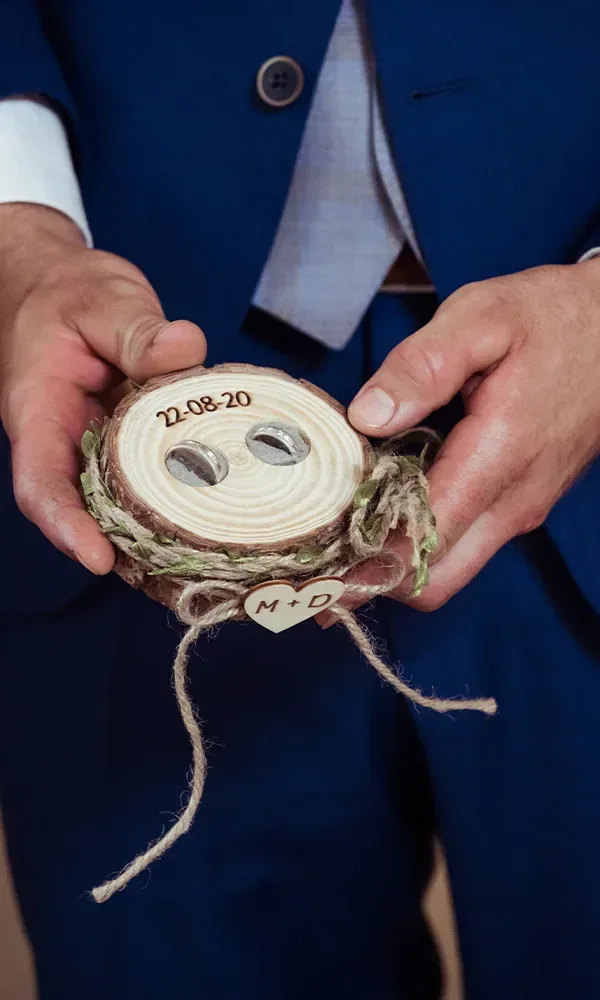 Der Bräutigam hält ein handgefertigtes Ringkissen aus Holz mit den Eheringen und dem Hochzeitsdatum '22.08.20'. Das Kissen ist mit Juteschnur und einem Herzanhänger mit den Initialen 'M+D' verziert.