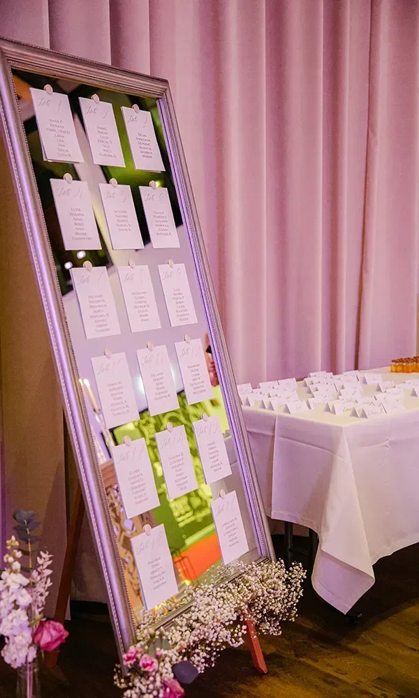 Ein eleganter Sitzplan für eine Hochzeit, präsentiert in einem großen, verspiegelten Rahmen. Auf dem Spiegel sind Karten mit den Namen der Gäste angebracht, die den jeweiligen Tischen zugewiesen sind. Der Rahmen ist mit Blumen am unteren Rand geschmückt, und im Hintergrund sind rosa Vorhänge sowie ein Tisch mit weiteren Namenskarten zu sehen.