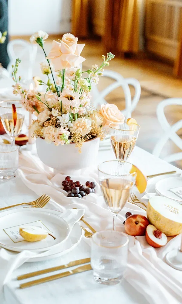 Ein festlich gedeckter Hochzeitstisch, dekoriert mit einem Blumenarrangement in Pastelltönen und frischem Obst wie Melone und Pfirsichen. Goldbesteck, Champagnergläser und elegante Geschirrteile vervollständigen die edle Atmosphäre.