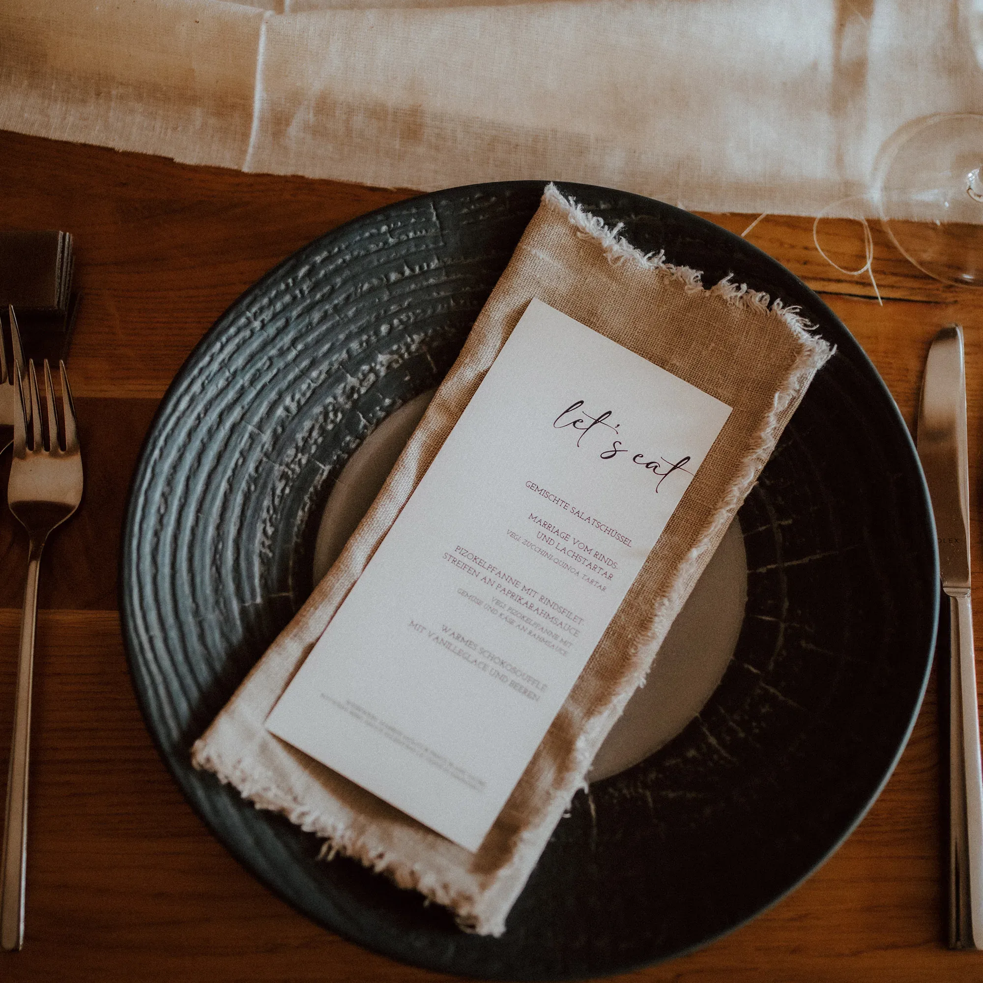 Ein detailreich gedeckter Hochzeitstisch mit einem dunkelgrauen Teller, darauf eine beige Serviette und eine handgeschriebene Menükarte mit der Aufschrift „Let’s eat“.