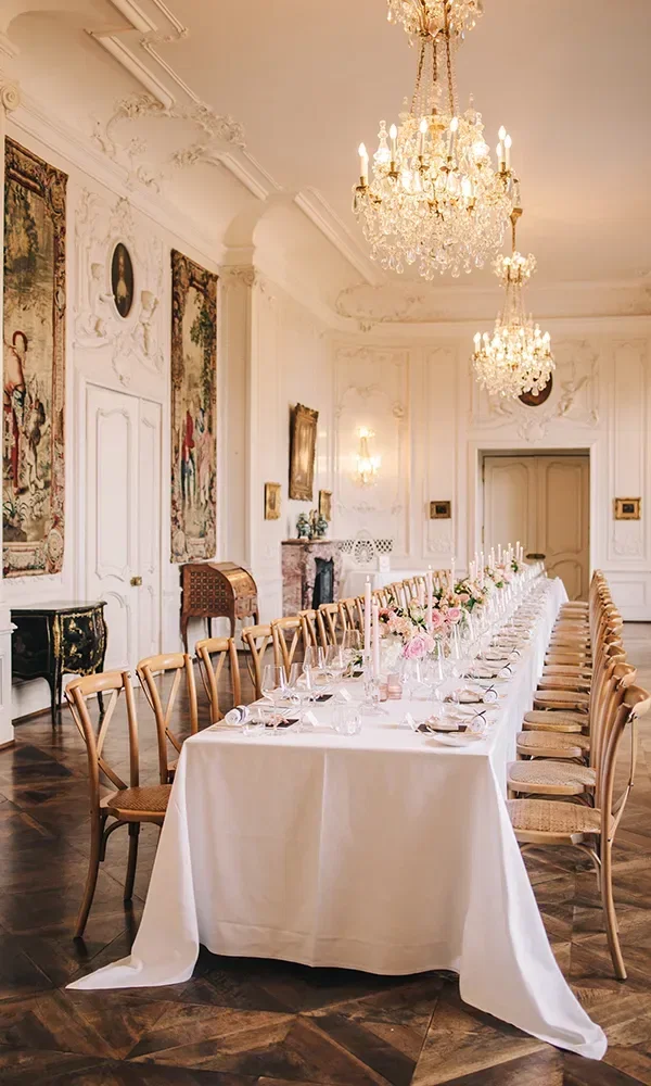 Eine lange, festlich gedeckte Tafel in einem prunkvollen Saal mit Kristalllüstern und Wandteppichen. Die Tischdekoration besteht aus eleganten Kerzen und Blumen.