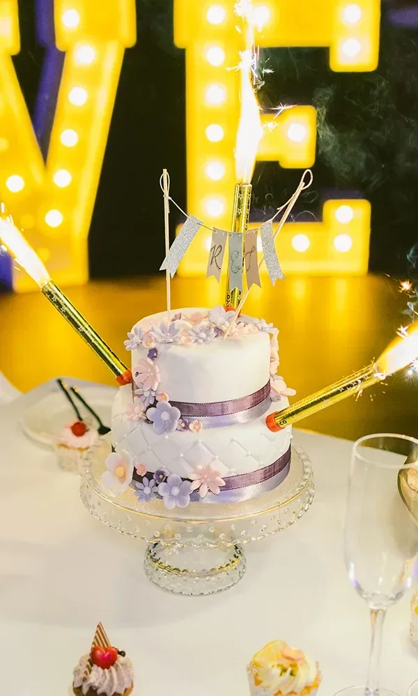 Eine wunderschön verzierte zweistöckige Hochzeitstorte mit lila Bändern und Blumen aus Zuckerguss. Die Torte ist auf einem gläsernen Tortenständer platziert, und funkelnde Wunderkerzen sind in die Torte gesteckt. Im Hintergrund leuchtet der Schriftzug 'LOVE' in gelben Buchstaben.