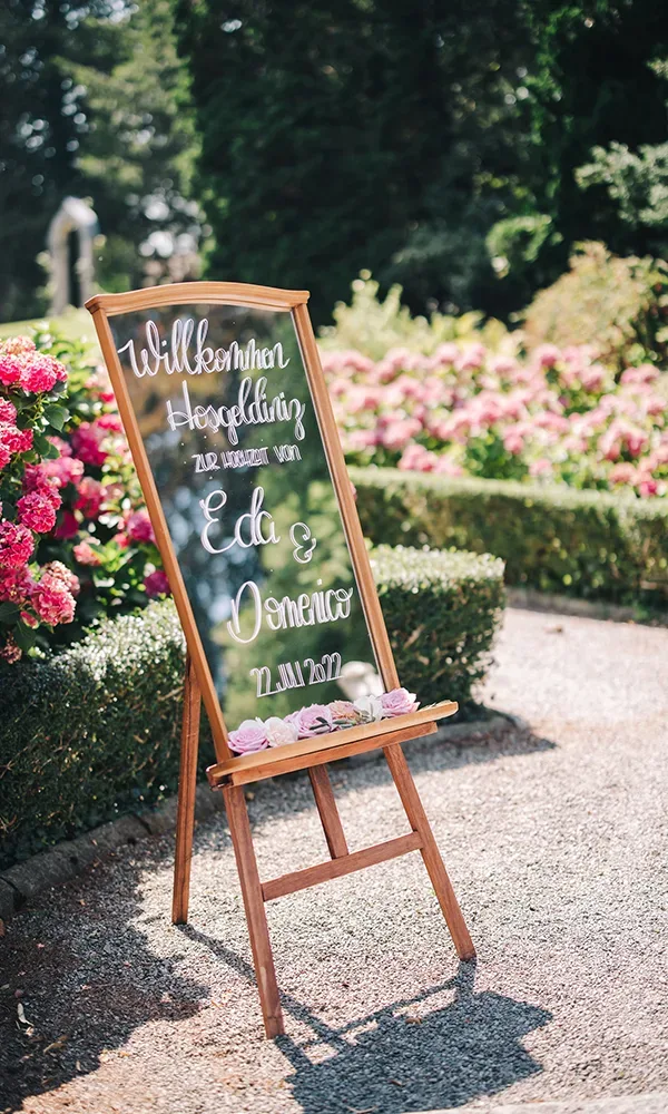 Ein hölzernes Willkommensschild mit den Namen des Brautpaares und dem Hochzeitsdatum, umgeben von üppiger Blumenpracht in einem Garten.