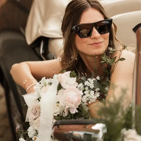 Eine lächelnde Frau mit stilvoller Sonnenbrille lehnt sich an ein Auto, während sie einen großen Strauß aus weißen und rosafarbenen Blumen hält. Ihr Haar ist locker gestylt, und sie trägt elegante Perlenarmbänder.