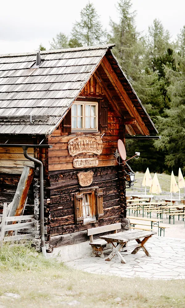 Eine rustikale Almhütte mit hölzerner Fassade und kleinen Fenstern, umgeben von Wald. Vor der Hütte stehen Holzbänke und Tische auf einem steinernen Untergrund.