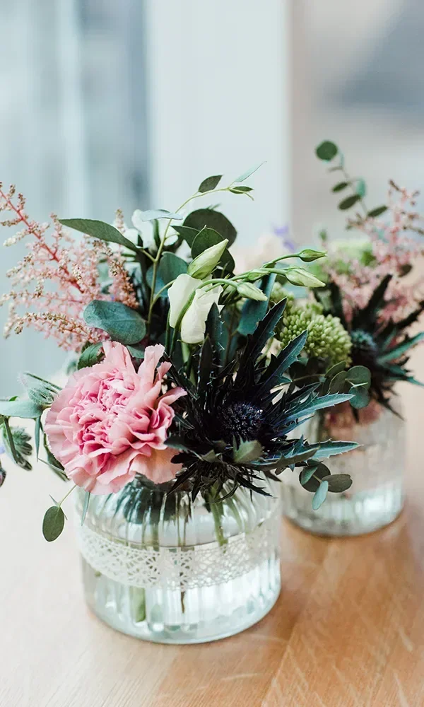Zwei Vasen mit farbenfrohen Blumengestecken, bestehend aus rosa, grünen und violetten Blüten, verziert mit Spitzenbändern, auf einem Holztisch arrangiert.