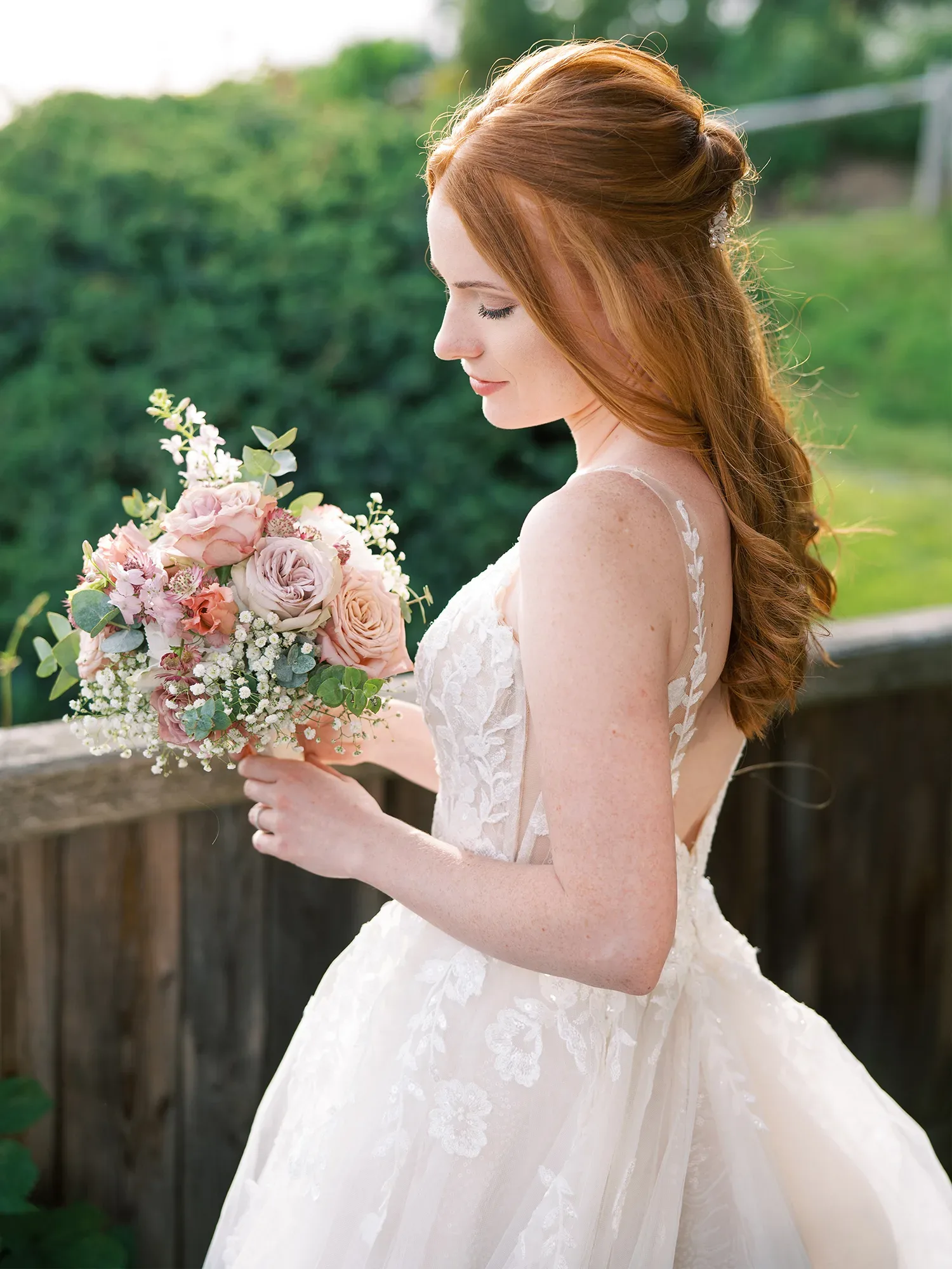 Profilaufnahme einer Braut mit langen roten Haaren, die einen Blumenstrauß mit rosafarbenen Rosen und Eukalyptusblättern hält, im Hintergrund grüne Landschaft.