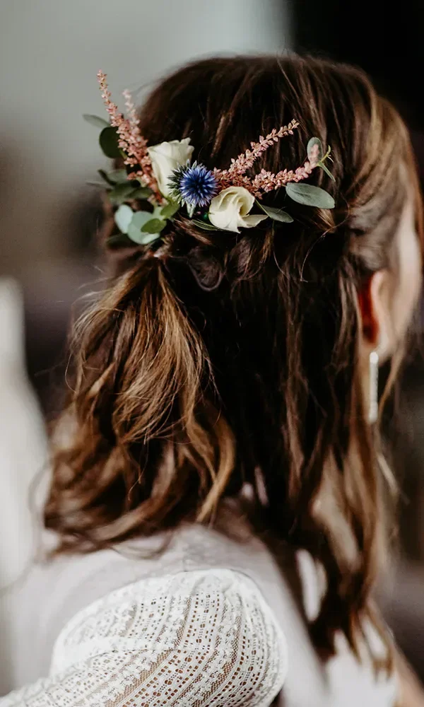 Braut mit halboffenem Haar und einem Blumenschmuck aus zarten, pastellfarbenen Blüten und grünen Blättern, elegant in die Frisur eingeflochten.