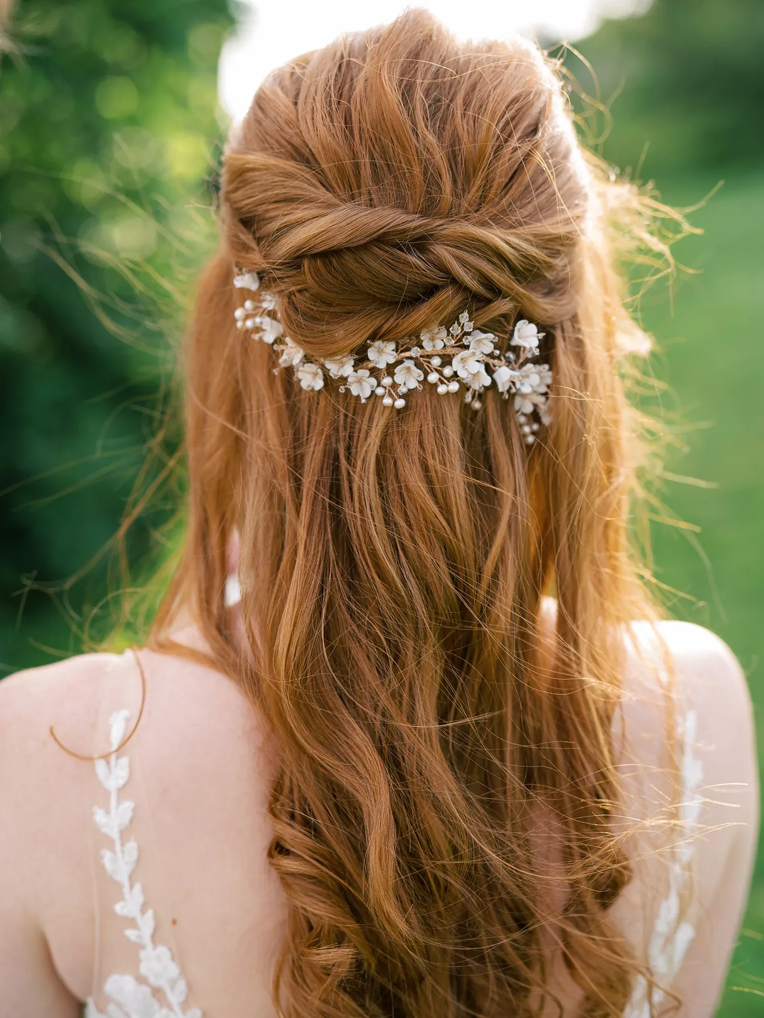 Eine Nahaufnahme der Rückenansicht einer Braut mit langen, gewellten roten Haaren, die zu einer halben Hochsteckfrisur mit weißen Blumenaccessoires gestylt sind.