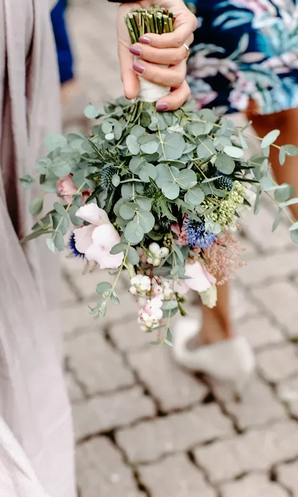 Ein wunderschöner Brautstrauß aus Eukalyptusblättern, zarten rosa und blauen Blumen, gehalten von der Braut mit frisch lackierten Fingernägeln.