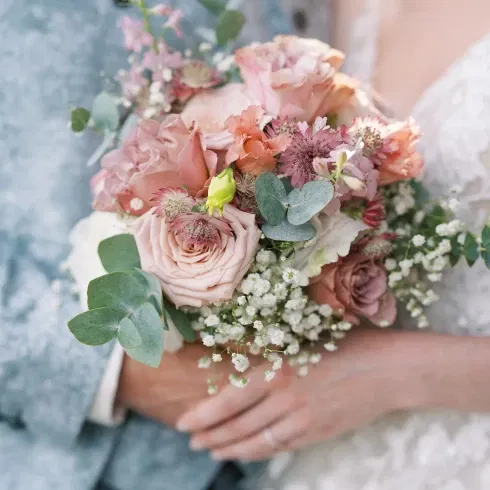 Nahaufnahme eines zarten Brautstraußes mit rosafarbenen Rosen, Eukalyptusblättern und Schleierkraut, gehalten von einem Brautpaar in eleganter Hochzeitskleidung.