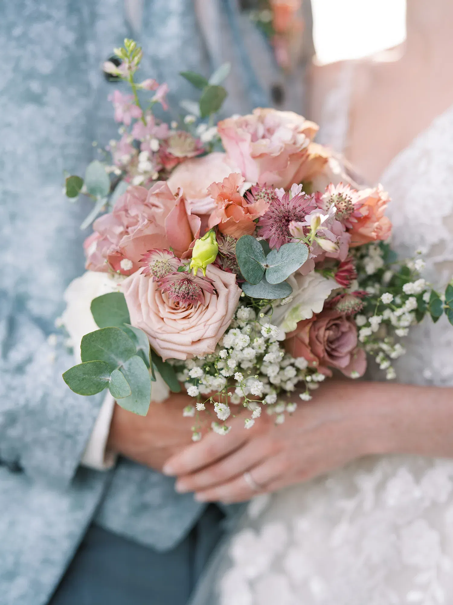 Nahaufnahme eines zarten Brautstraußes mit rosafarbenen Rosen, Eukalyptusblättern und Schleierkraut, gehalten von einem Brautpaar in eleganter Hochzeitskleidung.
