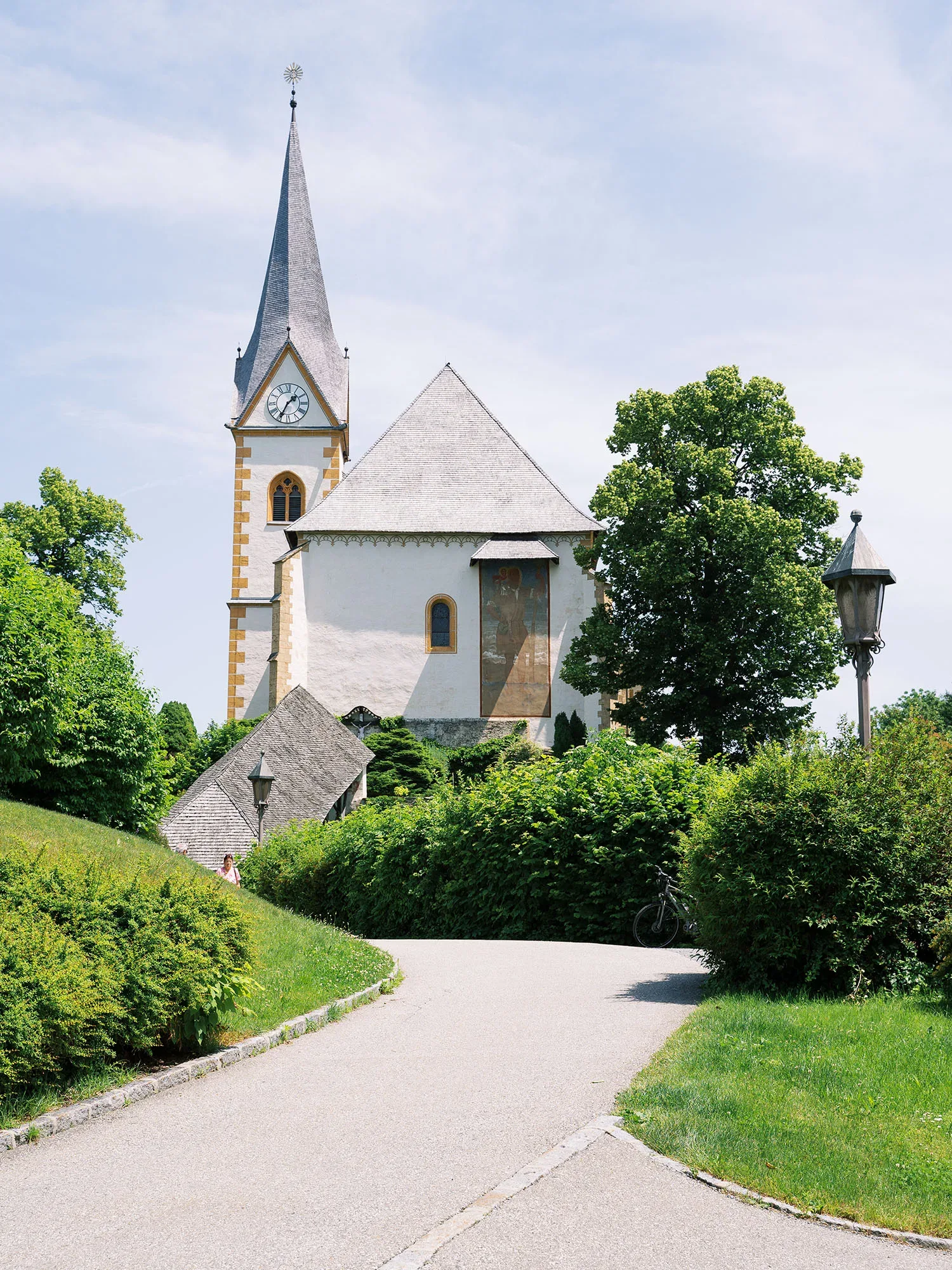 Eine malerische Kirche mit einem hohen Turm und einer Uhr, eingebettet in eine grüne, ruhige Landschaft mit einem gepflasterten Weg im Vordergrund.