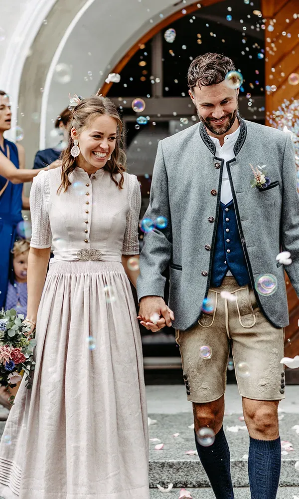 Braut und Bräutigam gehen Hand in Hand durch einen Bogen aus Seifenblasen. Sie trägt ein traditionelles Kleid, er eine Tracht mit Weste und Lederhose.