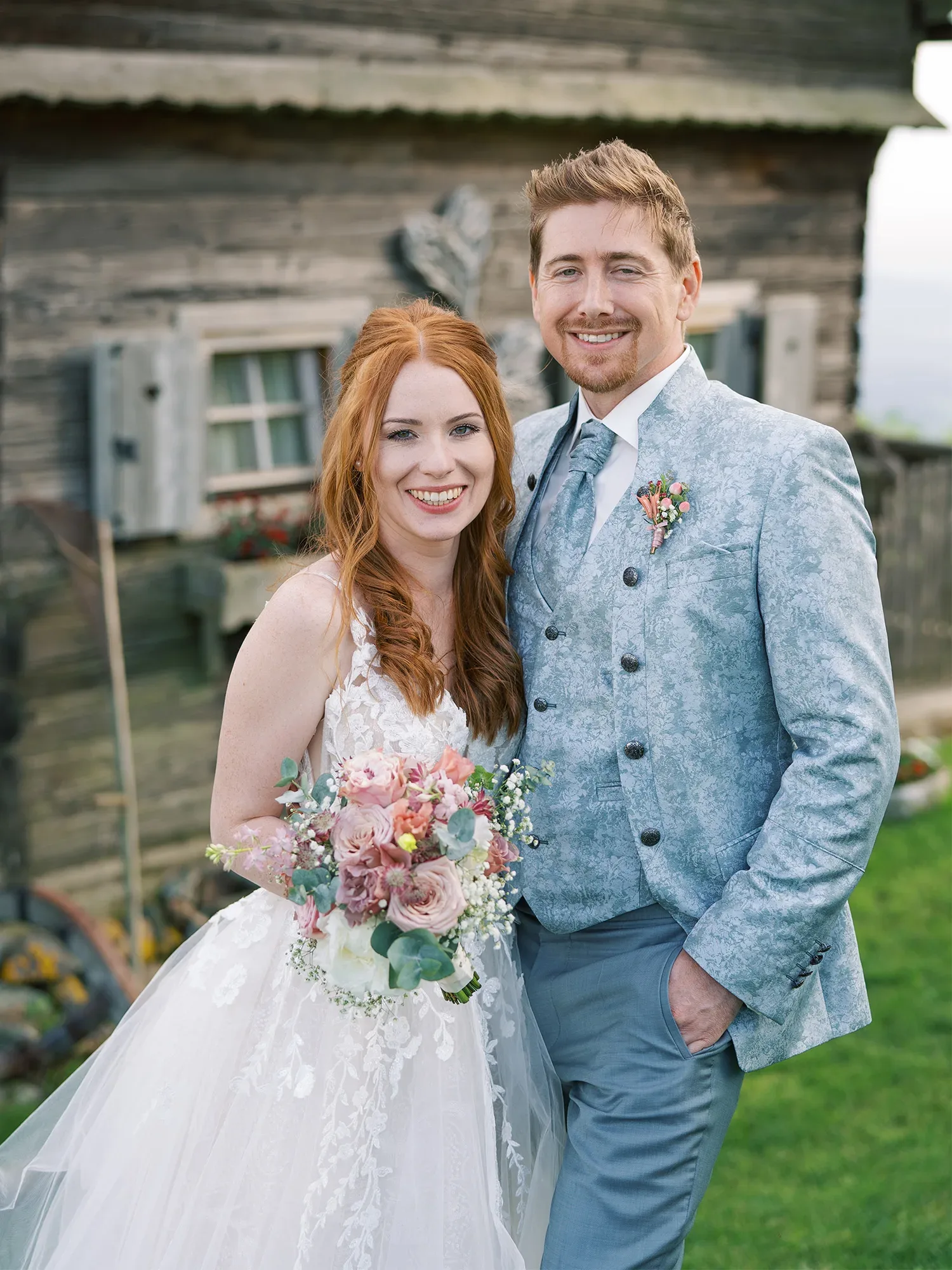 Hochzeitspaar lächelt vor einem rustikalen Holzgebäude, die Braut hält einen Blumenstrauß aus rosafarbenen und weißen Blumen, während der Bräutigam einen eleganten grauen Anzug trägt.
