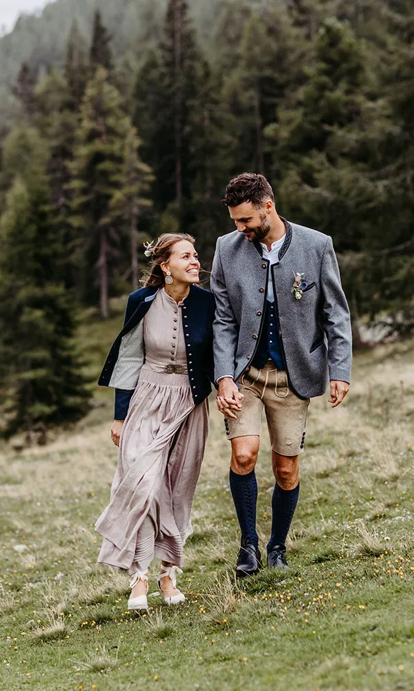 Ein Brautpaar in traditioneller Tracht, spazierend durch eine alpine Wiese. Die Braut trägt ein langes Kleid und der Bräutigam eine Lederhose mit Trachtenjacke. Beide lachen und halten sich an den Händen.