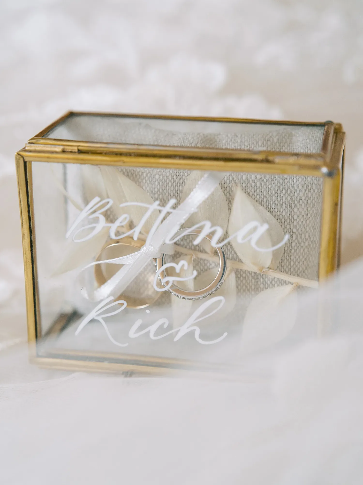 Zwei Eheringe in einer dekorativen Glasbox mit den Namen "Bettina & Rich", elegant arrangiert auf einem weißen Band.