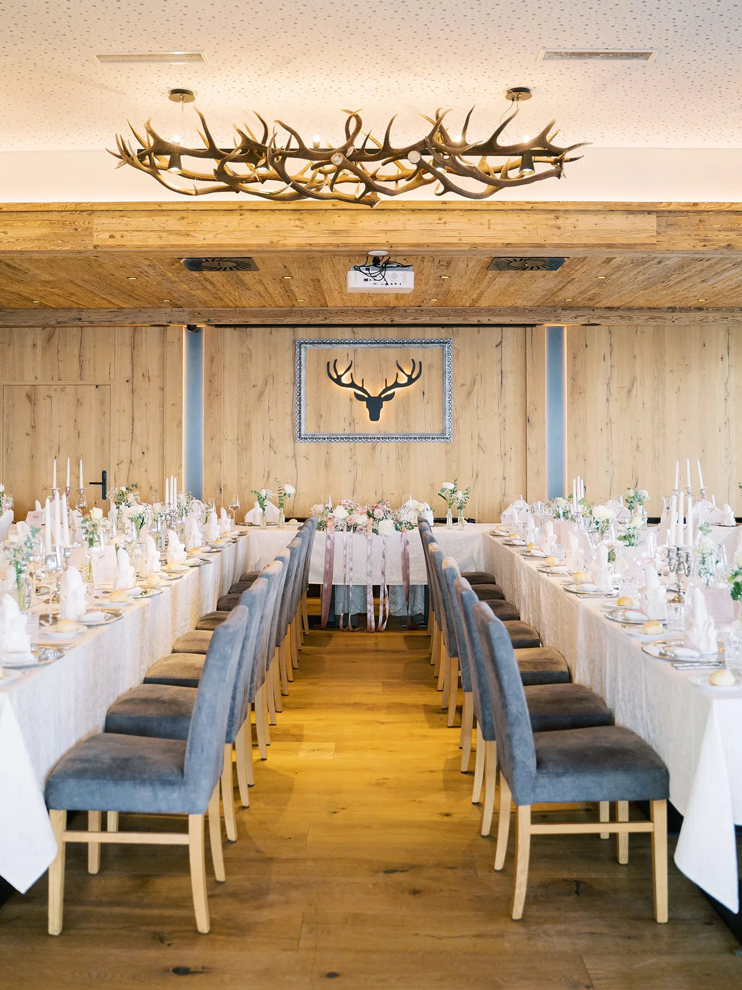 Festlich gedeckter Hochzeitssaal mit langen Tafeln und grauen Stühlen, im Hintergrund rustikale Holzwände und ein Hirschgeweih an der Wand.
