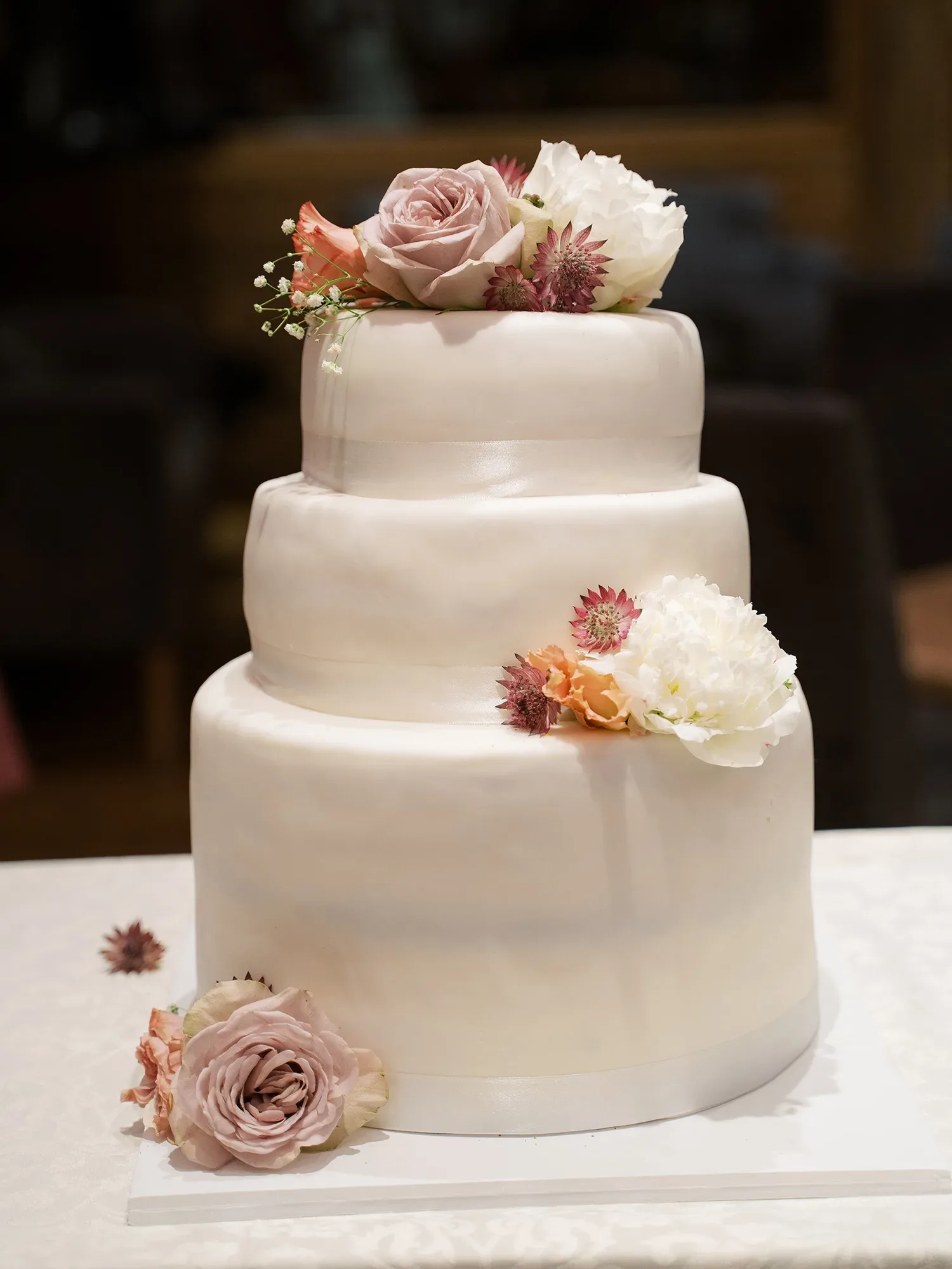 Dreistöckige, weiße Hochzeitstorte, dekoriert mit zarten Rosen und Blumen in Pastellfarben, steht auf einem weißen Tisch.
