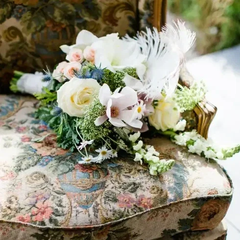 Ein eleganter Brautstrauß aus weißen und rosafarbenen Blumen liegt auf einem antiken Stuhl mit floraler Polsterung.