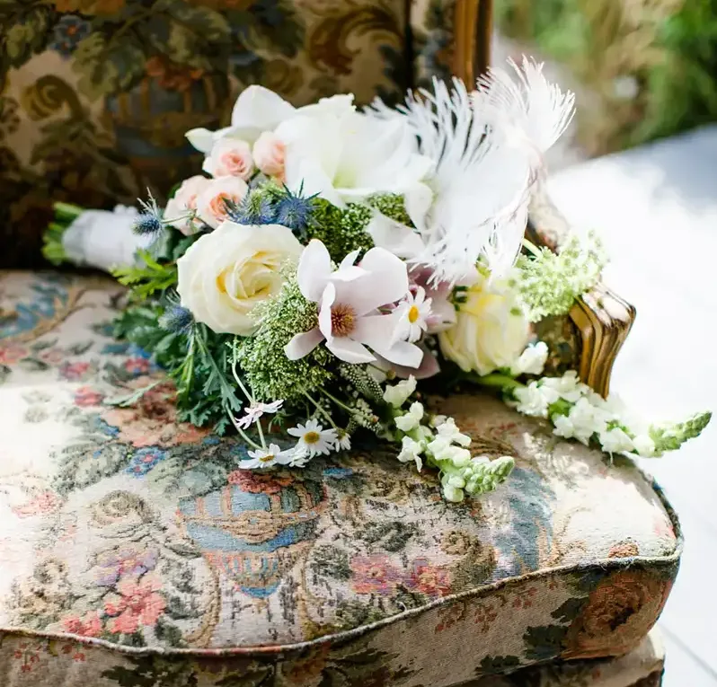 Ein eleganter Brautstrauß aus weißen und rosafarbenen Blumen liegt auf einem antiken Stuhl mit floraler Polsterung.