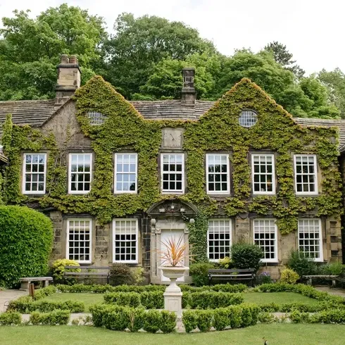 Ein wunderschönes historisches Haus, das mit Efeu bewachsen ist und eine symmetrische Fassade mit zahlreichen Fenstern und Steinwänden hat. Das Haus ist von einem gepflegten Garten umgeben, mit üppigem Grün, ordentlich geschnittenen Hecken, Bänken und einem zentralen Steinbrunnen. Das Anwesen liegt eingebettet in einer ruhigen, bewaldeten Gegend.