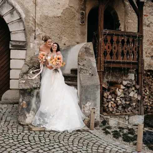 Zwei Bräute in traumhaften weißen Kleidern, posieren glücklich vor einer rustikalen Steinmauer.