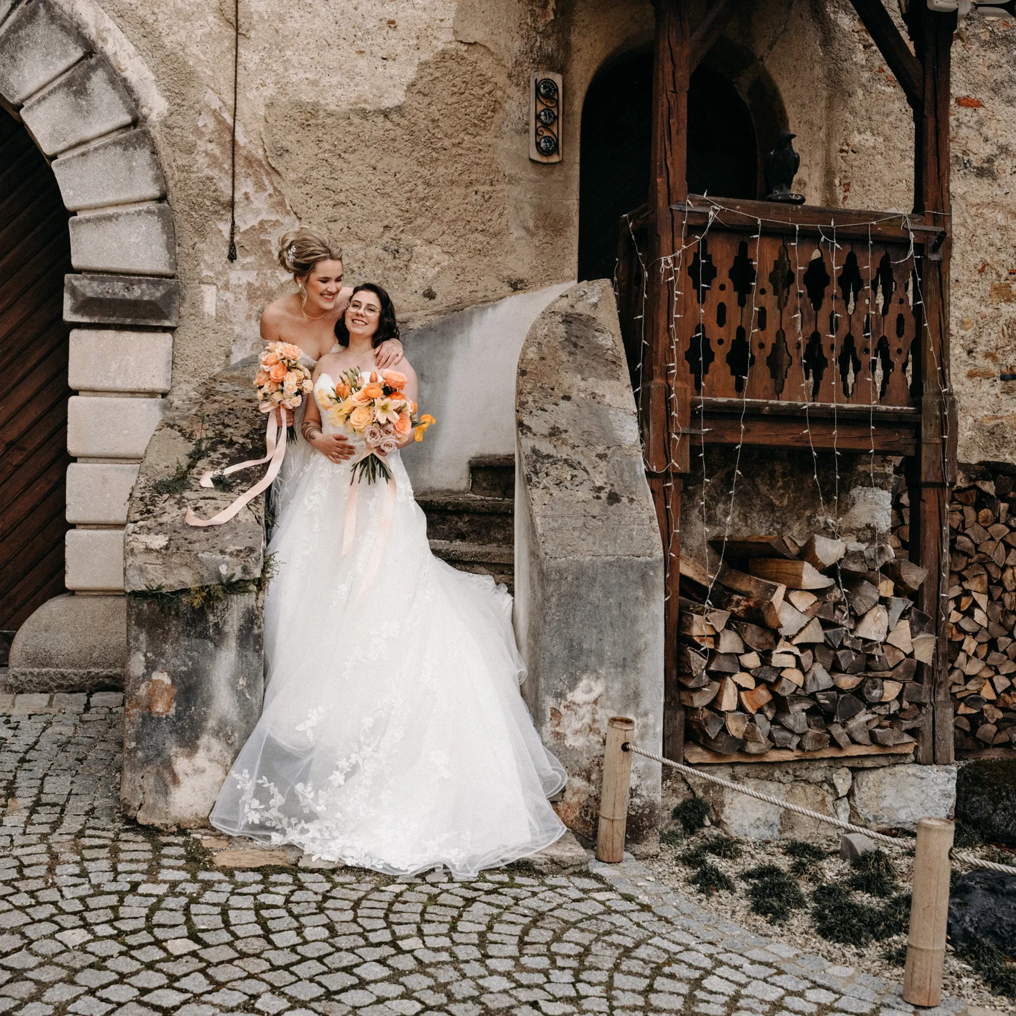 Zwei Bräute in traumhaften weißen Kleidern, posieren glücklich vor einer rustikalen Steinmauer.