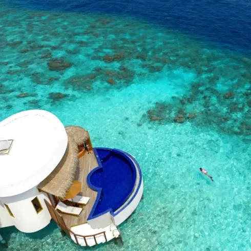 Honeymoon Ocean Suite mit einem blauen Infinity-Pool auf einer privaten Terrasse über dem kristallklaren, türkisfarbenen Wasser eines tropischen Riffs, mit einer Person, die im ruhigen Meer schwimmt.