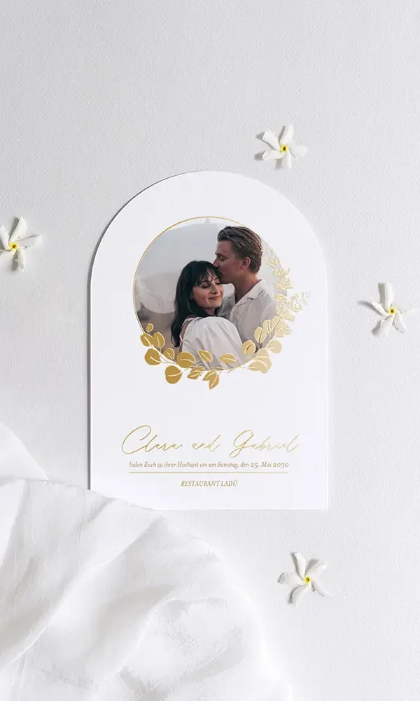 Eine elegante Hochzeitseinladung mit einem abgerundeten Design, die ein Bild eines lächelnden Paares umrahmt von einem goldenen floralen Muster zeigt. Unterhalb des Bildes stehen die Namen "Clara und Jakob" und eine Einladung zur Hochzeit am Samstag, den 25. Mai 2030 im Restaurant Lado.