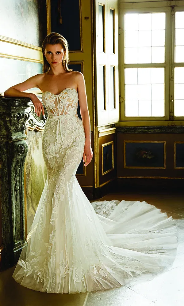 Eine Frau posiert in einem eleganten Hochzeitskleid vor einem klassischen Interieur. Das Kleid hat ein herzförmiges Korsett, das reich mit Blumenmustern bestickt ist, und geht über in einen vollen, spitzenbesetzten Meerjungfrauenrock mit einer ausladenden Schleppe. Das sanfte Licht von den Fenstern beleuchtet das Kleid und schafft eine stimmungsvolle und luxuriöse Atmosphäre.