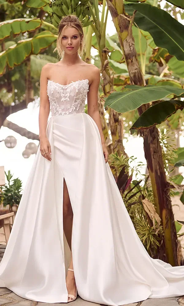 Eine Frau steht in einem tropischen Garten, gekleidet in ein stilvolles Hochzeitskleid mit einem trägerlosen Spitzenoberteil und einem glatten Satinrock mit einem hohen Schlitz vorne. Das Kleid hat eine A-Linien-Silhouette mit einer fließenden Schleppe, die Eleganz und moderne Raffinesse ausstrahlt.