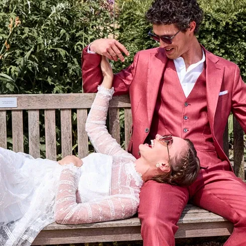 Lächelndes Brautpaar, die Braut in einem Spitzenkleid und der Bräutigam in einem roten Anzug, entspannt auf einer Bank im Garten.