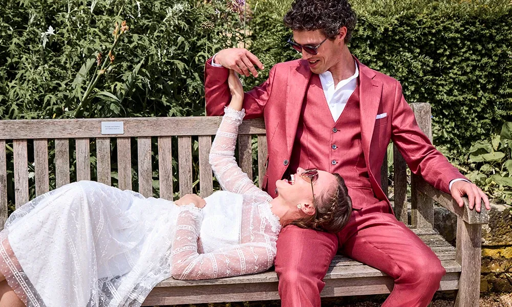 Lächelndes Brautpaar, die Braut in einem Spitzenkleid und der Bräutigam in einem roten Anzug, entspannt auf einer Bank im Garten.