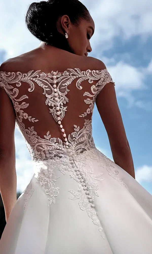 Ein Blick auf den Rücken einer Frau, die ein hochgeschlossenes Hochzeitskleid mit einer detaillierten, durchscheinenden Spitzenrückseite trägt, die mit aufwendigen Stickereien und Perlenknopfverschlüssen verziert ist. Das Kleid zeichnet sich durch eine fließende Silhouette aus und die Perspektive gegen den blauen Himmel betont die exquisite Handwerkskunst und die romantischen Details des Kleides.