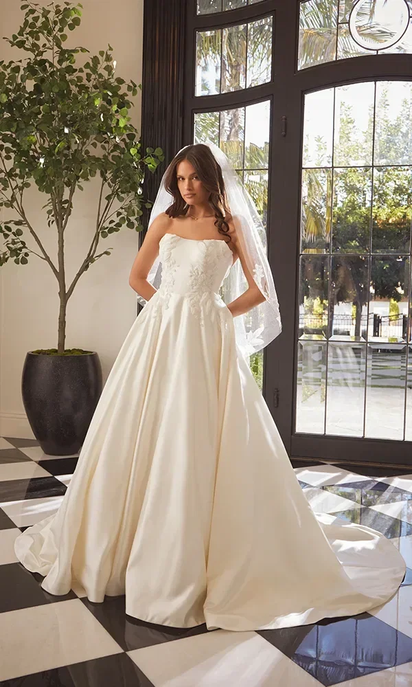 Hier steht die Braut vor einer Tür mit Glasscheiben und Blick auf Palmen, in einem romantischen Kleid mit Spitzenoberteil und langer Schleppe. Der transparente Schleier fällt sanft um ihre Schultern.