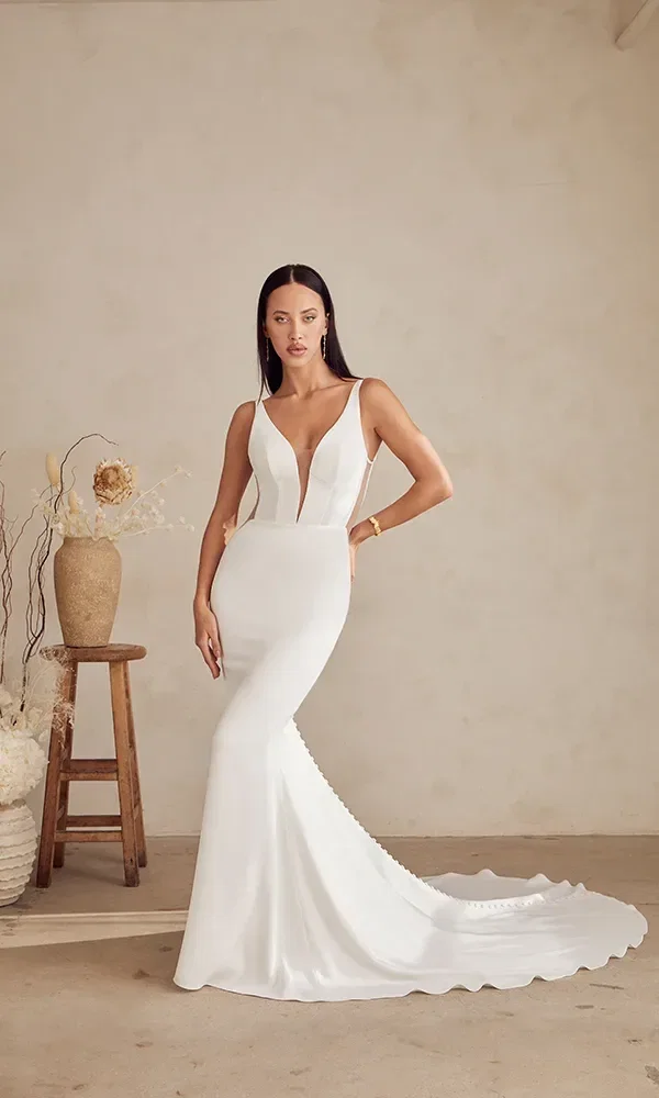 Das Bild zeigt eine Braut in einem schlichten, aber eleganten figurbetonten Kleid mit tiefem V-Ausschnitt. Sie steht neben einem rustikalen Holzhocker und trockenen Pflanzen, was einen kontrastreichen Hintergrund zu ihrem modernen Kleid bildet.