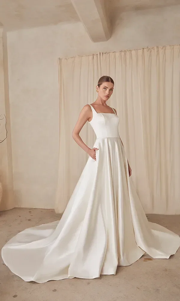 Die Braut in einem minimalistischen Brautkleid mit einem fließenden Rock und einer langen Schleppe posiert stehend in einem Raum mit neutralem Hintergrund, was ihre elegante Statur unterstreicht.