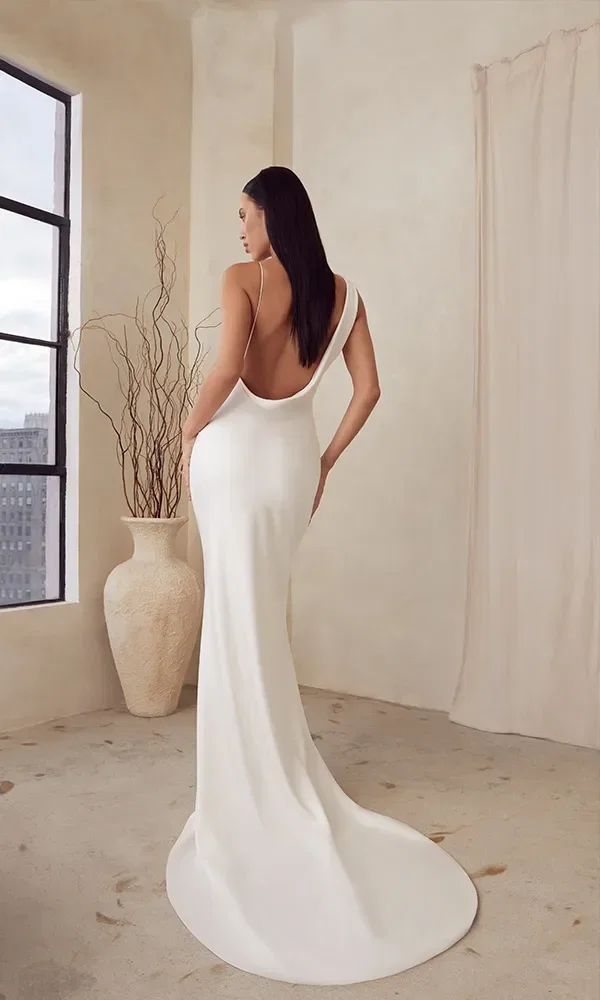 Rückansicht einer Braut in einem figurbetonten Kleid mit einer eleganten niedrigen Rückenausschnitt. Sie steht in einem Raum mit einem großen Fenster, das Tageslicht hereinlässt.