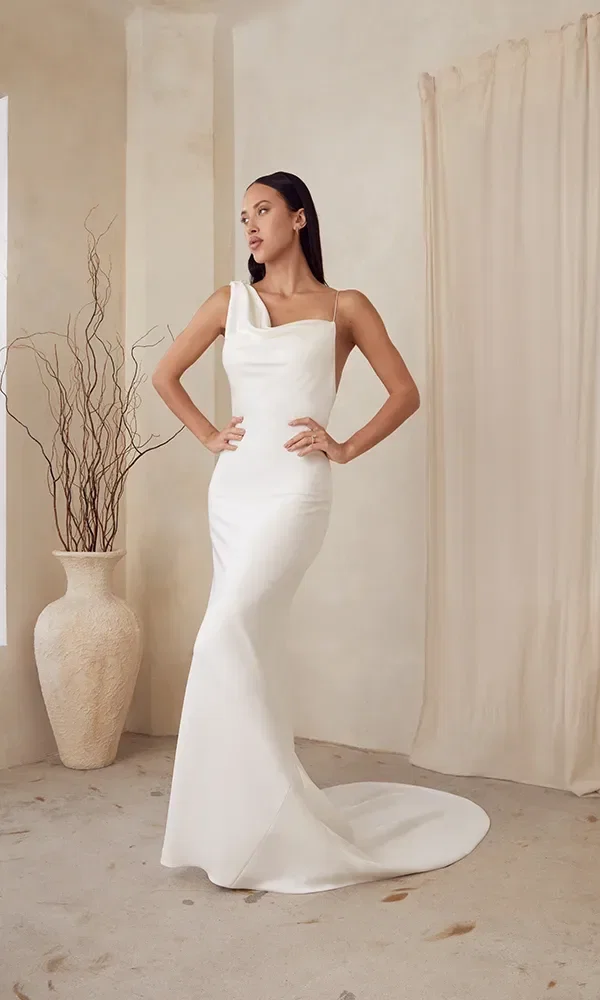 Eine Braut in einem Brautkleid mit einer hohen Beinschlitze und einem aufwendigen Spitzenoberteil posiert im Innenraum mit direktem Blick auf einen sonnendurchfluteten Garten.