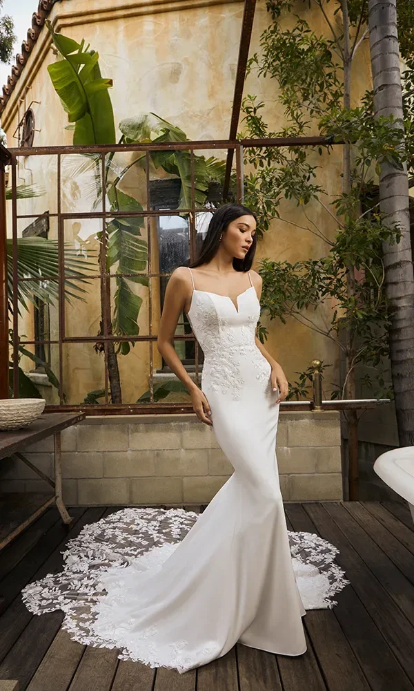 Eine Braut steht in einem naturnahen Innenhof, umgeben von grünen Pflanzen und einer alten, verwitterten Wand. Ihr Kleid ist schmal und fließend mit einer eleganten Spitzenverzierung, die an der Taille beginnt und sich über die Hüften erstreckt, mit einer dramatischen Schleppe, die mit Spitze besetzt ist.