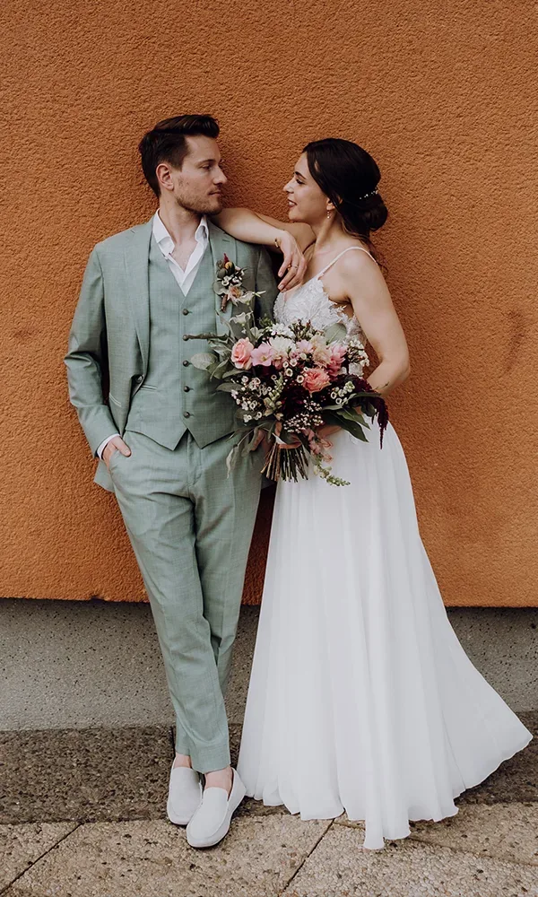 Ein elegantes Brautpaar steht vor einer orange-farbenen Wand. Der Bräutigam trägt einen mintgrünen Anzug und weiße Schuhe, die Braut ein schlichtes weißes Kleid und hält einen Strauß mit rosa und weißen Blumen.
