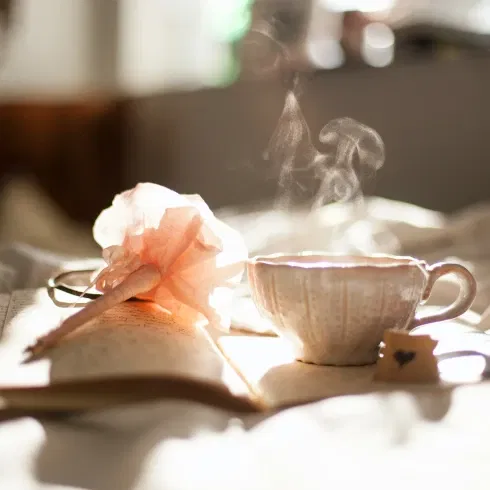 Eine Dampfende Teetasse steht auf einem aufgeklappten Buch, daneben ein Stift mit Blume