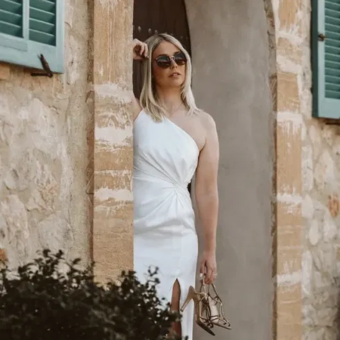 Eine stilvolle Frau in einem weißen, schulterfreien Kleid lehnt lässig an einer Steinmauer und hält ihre goldenen Sandaletten in der Hand. Sie trägt Sonnenbrillen und posiert vor einem rustikalen Gebäude mit grünen Fensterläden.