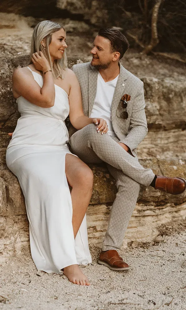 Ein verliebtes Paar sitzt eng beieinander auf einer Steinmauer. Die Frau in einem ärmellosen weißen Kleid blickt den lächelnden Mann an, der einen grauen Tweed-Blazer, ein weißes Hemd und braune Lederschuhe trägt, seine Sonnenbrille steckt im Blazer.