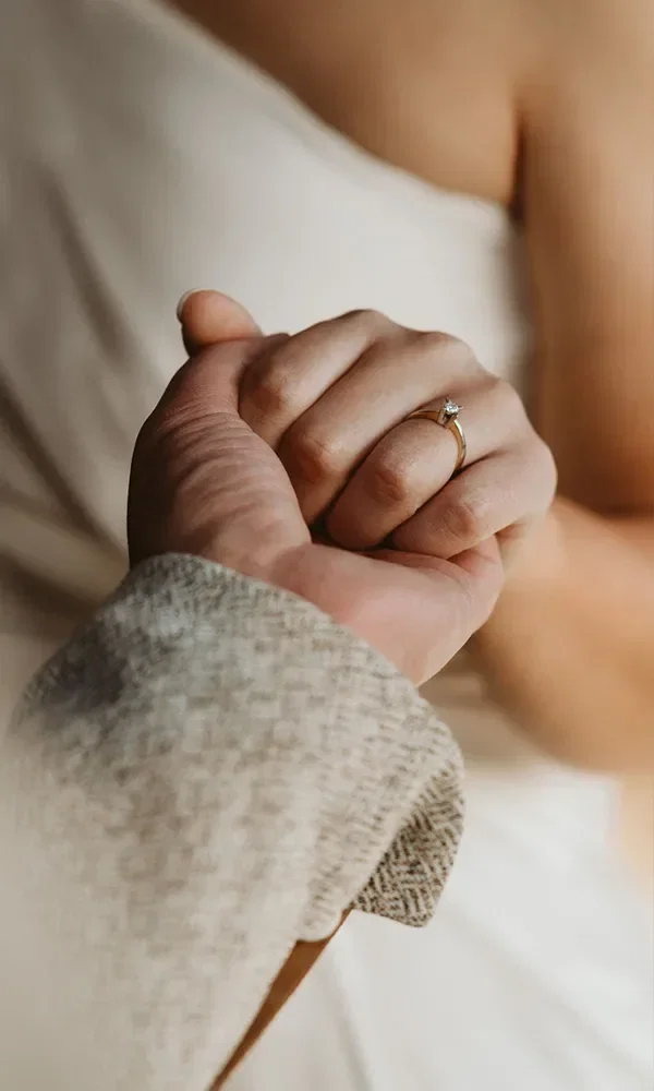 Die Hand der Frau liegt in der des Mannes, ein schlichter Verlobungsring mit einem einzelnen Diamanten auf einem goldenen Band ist am Finger der Frau zu sehen.