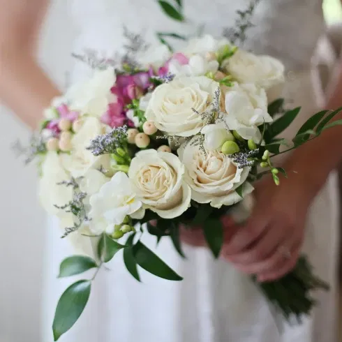 Eine Frau in einem weißen Hochzeitskleid hält einen Braustrauß aus weißen Rosen mit vereinzelt rosafarbenen Blumen in der Hand