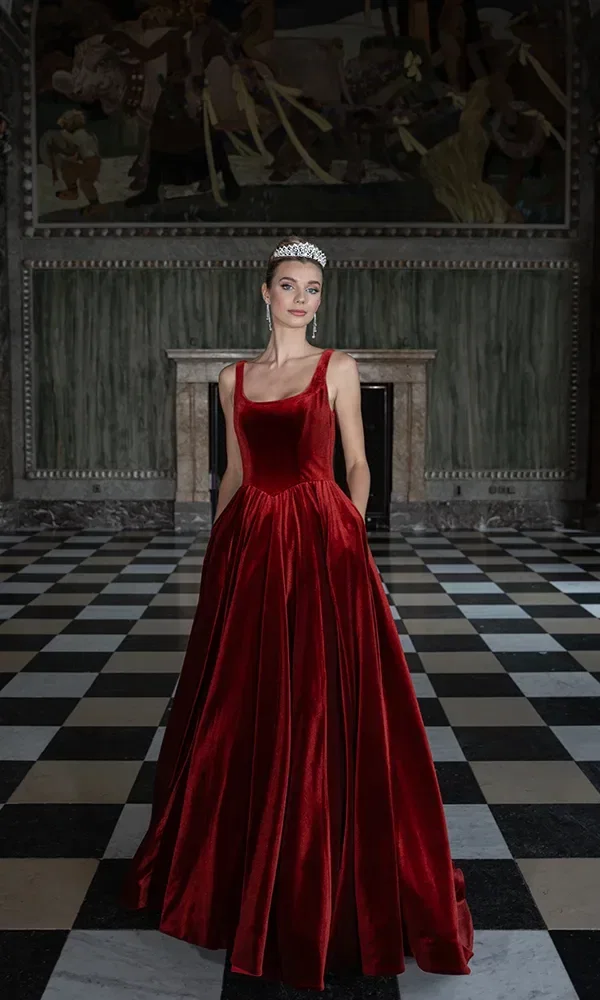 Eine Frau in einem tiefroten Samtkleid mit V-Ausschnitt steht selbstbewusst vor einem historischen Wandgemälde und einem schachbrettartigen Boden, gekrönt mit einem funkelnden Diadem.