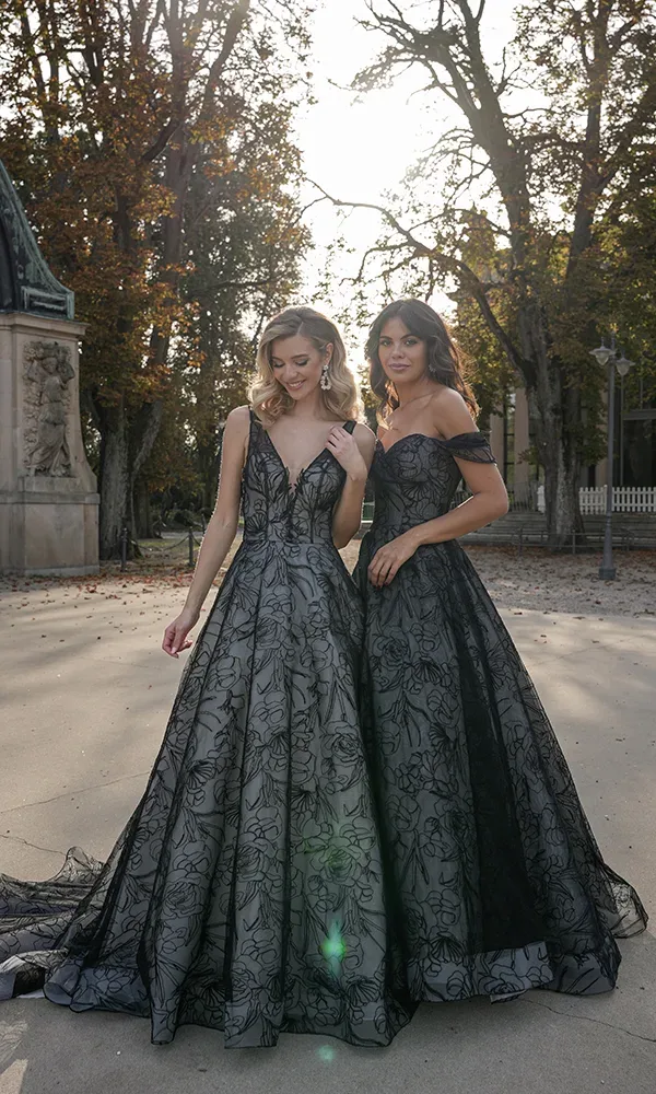 Zwei Frauen in eleganten, bodenlangen Abendkleidern mit Spitzen-Overlay stehen nebeneinander auf einem Gehweg mit herbstlichen Bäumen im Hintergrund, die Sonne scheint durch die Blätter.