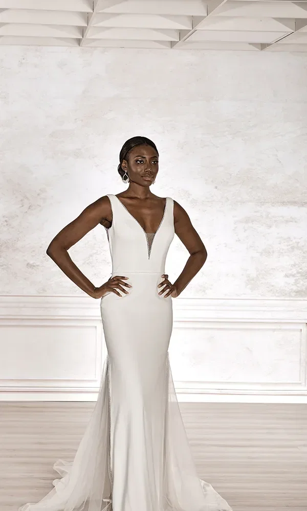 Eine Frau in einem weißen, figurbetonten Brautkleid mit V-Ausschnitt und strassbesetztem Dekolleté steht vor einem hellen Hintergrund, die Hände elegant auf die Hüften gelegt.